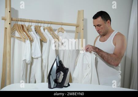 Blanchisserie nettoyage livraison de nourriture divers travaux ménagers hommes un homme et un fer dans ses mains vêtements qui ont besoin d'être repassés cintre en bois fer planche à repasser confort dans la maison est son propre patron homme mari pendant une heure Banque D'Images