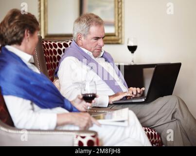Se détendre dans la soirée. Un homme âgé sur son ordinateur portable tout en prenant un verre avec sa femme. Banque D'Images