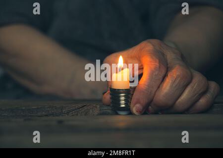 La vieille main d'une femme tient une bougie dans une cartouche d'une ampoule sur une table à la maison dans l'obscurité sans lumière, une attaque de missile en Ukraine, les gens W. Banque D'Images