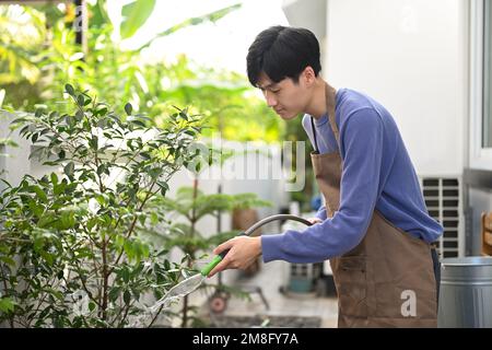Joyeux jeune homme asiatique dans le tablier arroser les plantes du tuyau à l'arrière-cour. Jardinage concept de passe-temps Banque D'Images