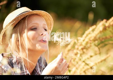 Amusement et fantaisie dans les champs. Belle jeune femme portant une fedora de paille et soufflant à un pissenlit. Banque D'Images