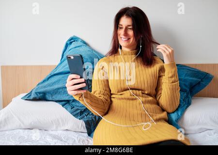 Femme avec écouteurs souriant lors d'un appel vidéo sur le téléphone mobile dans le lit. Banque D'Images