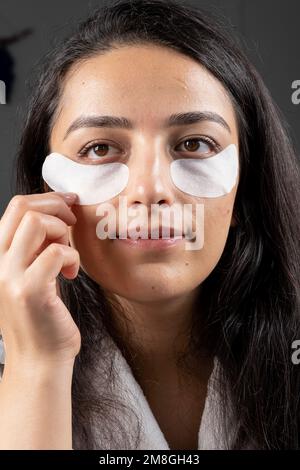 Sous les timbres oculaires, fermez la femme brunette millénaire caucasienne qui applique sous les timbres oculaires. Miroir de recherche utilisant la routine de soin cosmétique anti-âge Banque D'Images