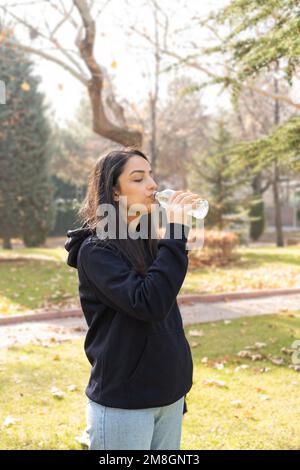 Femme assoiffée de boire de l'eau à partir d'une bouteille en verre. En plein air au parc. Ensoleillé mais froid. Feuilles jaunes tombées sur l'herbe. Copier l'espace. Banque D'Images