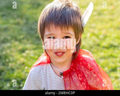 Portrait d'un garçon souriant jouant indien américain. Enfant avec plume d'oiseau blanche et manteau rouge. Jeu de rôle de costume. Activités de loisirs en plein air. Banque D'Images