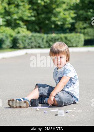 Un petit enfant attentif est assis sur la chaussée dans le parc. Kid dessine quelque chose sur l'asphalte. Activités de loisirs créatives pour les enfants. Banque D'Images