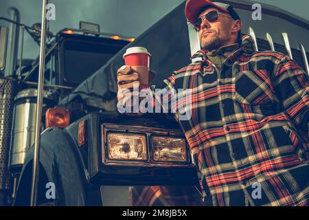 American semi Trucker chauffeur prenant une courte pause sur un arrêt de camion. Boire du café devant son véhicule. Homme caucasien dans son 40s. Banque D'Images