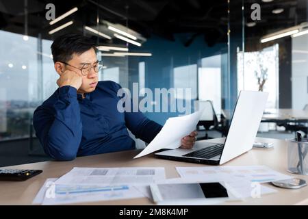 Jeune asiatique fatigué, comptable, homme d'affaires, indépendant. Il est assis au bureau à une table avec un ordinateur portable, regarde à travers les documents, tient sa tête dans ses mains, soupire une journée difficile et ennuyeuse. Banque D'Images