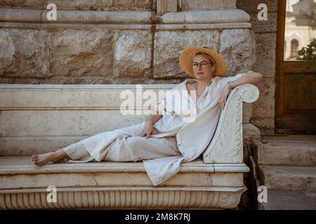 une belle femme mûre est assise sur un banc de pierre dans un chapeau et repose Banque D'Images