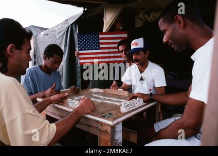 Un groupe de réfugiés migrants cubains jouent un jeu de dominos pour aider à passer le temps, en attendant de réaliser leur rêve « américain » dans le camp de Safe Haven 2, situé à Guantanamo Bay, Cuba. En arrière-plan, un réfugié détient un drapeau américain. De L'ARTICLE de mai 1995 du MAGAZINE AIRMAN intitulé « Trail of Dreams ». Base: Guantanamo Bay pays: Cuba (CUB) Banque D'Images