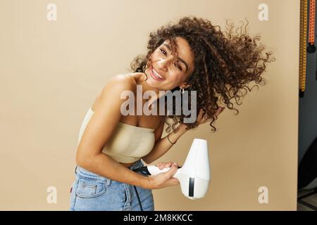 Femme sèche les cheveux freux avec sèche-cheveux, maison beauté produits coiffants cheveux, sourire sur fond beige Banque D'Images