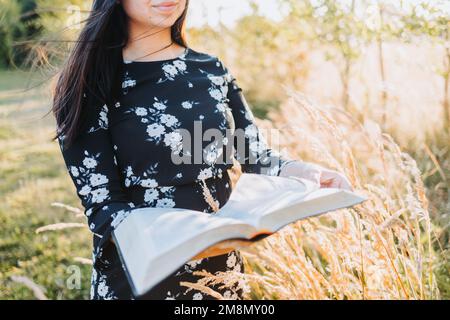 Jeune fille chrétienne religieuse tenant et lisant sa bible, à l'extérieur dans le champ au coucher du soleil. Communion avec Dieu. Banque D'Images