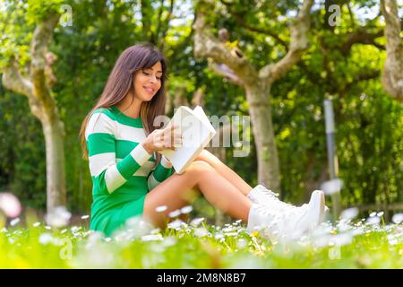 Portrait d'une fille de brune caucasienne lisant un livre dans la nature assis sur l'herbe portant un chandail vert et blanc Banque D'Images