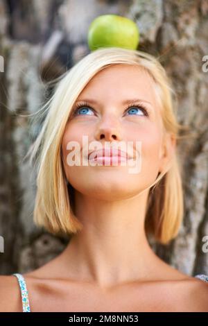 Rester en bonne santé nécessite un mode de vie équilibré. Jolie jeune femme penchée contre un tronc d'arbre avec une pomme équilibrée sur sa tête. Banque D'Images