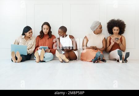 Groupe de cinq femmes d'affaires assises sur le sol contre un mur dans un bureau et utilisant la technologie. Les gens d'affaires et les collègues parlent et utilisent Banque D'Images