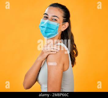 Race mixte Covid vaccinée femme montrant du plâtre sur le bras, portant un masque chirurgical. Portrait d'une femme hispanique isolée contre un studio jaune Banque D'Images