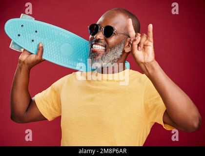 Un homme afro-américain mature debout avec un mini-skateboard en studio isolé sur fond rouge. Beau et insouciant homme de porter Banque D'Images