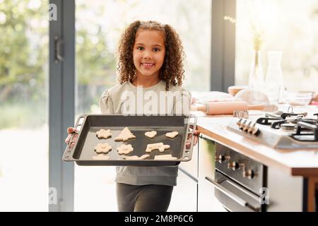 Une adorable petite fille de race mixte à l'air heureuse et fière tout en cuisant seul à la maison. Enfant hispanique avec des cheveux bouclés faisant des biscuits dans une cuisine Banque D'Images