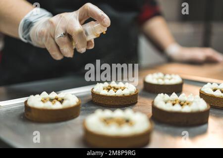 Boulanger femme professionnelle portant des gants et un tablier noir pulvérisant des paillettes comestibles dorées sur des tartes au chocolat à dessert mini. Processus de cuisson. Décoration. Gros plan horizontal en intérieur. Photo de haute qualité Banque D'Images