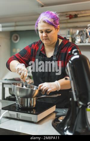 Boulanger femme professionnelle avec cheveux teints violets portant un tablier noir faisant de la ganache de chocolat dans le bain d'eau. Processus de cuisson. Desserts. Prise de vue verticale en intérieur. Photo de haute qualité Banque D'Images