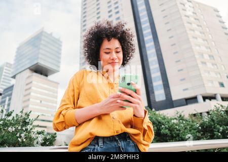 Jeune femme hispanique heureuse regardant des vidéos amusantes sur une application de médias sociaux pour smartphone en plein air. Latino dame avec chemise jaune SMS avec téléphone cellulaire dans la rue. Photo de haute qualité Banque D'Images
