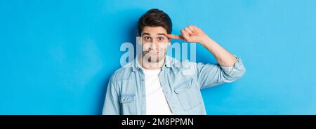 Gros plan d'un homme qui se colle pour agir stupide ou fou, le doigt sur la tête et regardant la caméra, debout sur fond bleu Banque D'Images