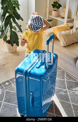 Enfant utilisant un téléphone portable achetant des billets en ligne ou attendant des vacances à la maison à côté d'une valise emballée Banque D'Images