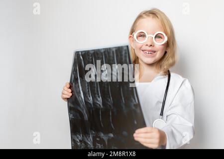 Enfant caucasien, petit médecin avec lunettes et stéthoscope, tenant film radiographique, scanner, IRM. Enfant souriant et regardant l'appareil photo allumé Banque D'Images