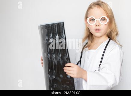 Enfant caucasien sérieux, petit médecin avec lunettes et stéthoscope jouet, tenant un film radiographique, scanner, IRM. Enfant regardant l'appareil photo allumé Banque D'Images