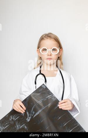 Enfant caucasien vêtu d'un uniforme de médecin avec des lunettes et un stéthoscope, qui a l'air triste et sérieux. Enfant tenant un film radiographique d'acquisition ct Banque D'Images