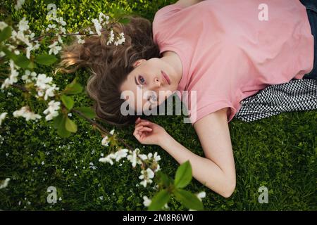 Belle femme long cheveux repose sur l'herbe dans les fleurs, regarde l'appareil photo. Modèle femelle au printemps Banque D'Images