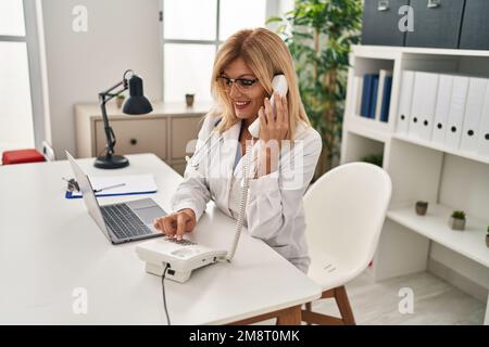 Femme blonde d'âge moyen portant l'uniforme du médecin parlant au téléphone à la clinique Banque D'Images