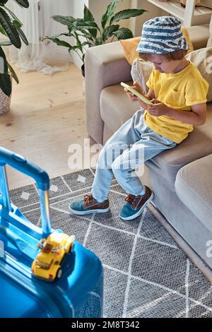 Enfant utilisant un téléphone portable achetant des billets en ligne ou attendant des vacances à la maison à côté d'une valise emballée Banque D'Images