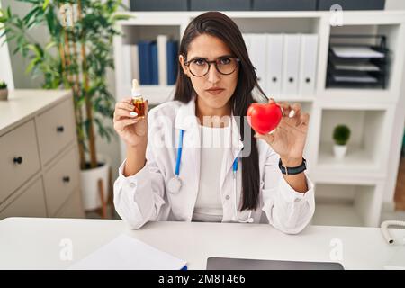 Jeune femme hispanique médecin tenant le coeur et le cbd huile sceptique et nerveux, frowning bouleversé à cause du problème. Personne négative. Banque D'Images