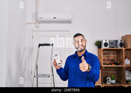 Réparateur hispanique travaillant avec le climatiseur souriant heureux et positif, pouce vers le haut faisant excellent et signe d'approbation Banque D'Images