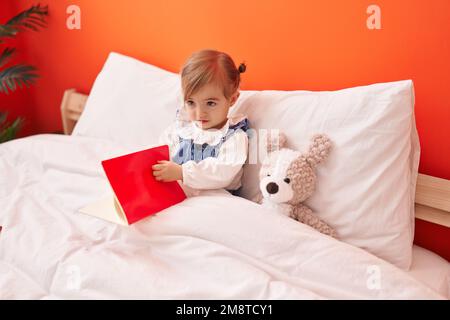 Adorable livre de lecture blond pour tout-petit assis sur le lit avec un ours en peluche dans la chambre Banque D'Images