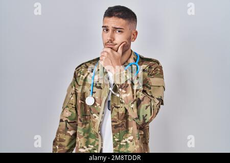 Jeune médecin hispanique portant un uniforme militaire de camouflage pensant concentré sur le doute avec le doigt sur le menton et regardant vers le haut se demander Banque D'Images