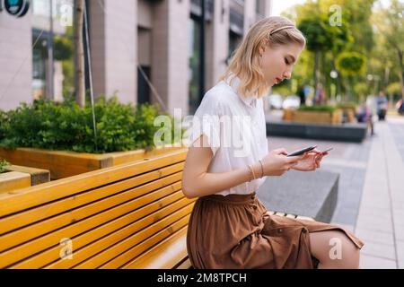 Vue latérale d'une belle jeune femme blonde tenant une carte de crédit en utilisant le téléphone mobile shopping en ligne, en choisissant quelque chose assis sur le banc sur la rue de la ville. Banque D'Images