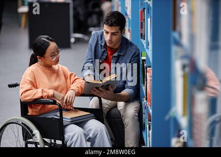 Portrait de la jeune femme asiatique en fauteuil roulant dans la bibliothèque et en choisissant des livres avec l'aide d'amis Banque D'Images