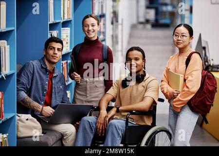 Groupe diversifié d'étudiants avec un jeune homme en fauteuil roulant regardant la caméra dans la bibliothèque de l'université, concept d'inclusion Banque D'Images