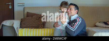 Image d'un père souriant embrassant sa fille en jouant à la maison et en chantant du karaoké. Bannière horizontale Banque D'Images