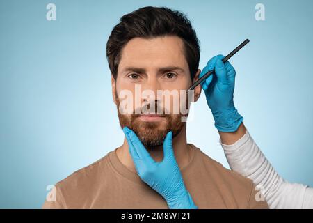 Le médecin dessine des marques sur le visage masculin pour une opération de chirurgie esthétique sur fond bleu de studio Banque D'Images