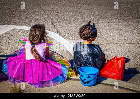 Deux enfants un garçon en costume de chauve-souris et une fille en costume de princesse arc-en-ciel s'assoient sur le trottoir en attendant le défilé d'Halloween Banque D'Images