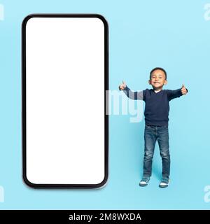 Adorable enfant afro-américain debout près d'un grand smartphone avec écran blanc et pouces Banque D'Images