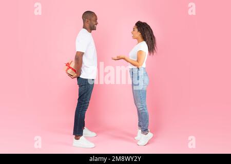 africain américain d'âge moyen homme cachant boîte cadeau derrière le dos et regardant la femme heureuse intéressée, fond rose Banque D'Images