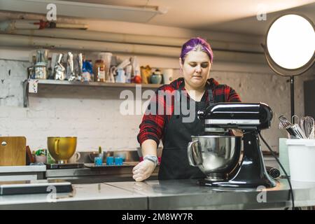 Boulanger femme professionnelle avec cheveux violets portant un tablier noir faisant de la crème de beurre givrant dans le batteur sur pied. Cuisine professionnelle. Processus de cuisson. Prise de vue horizontale en intérieur. Photo de haute qualité Banque D'Images