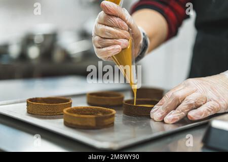 Vue rapprochée d'un boulanger portant des gants pour le remplissage de caramel dans des mini-croûtes vides. Processus de cuisson professionnel. Assemblage du dessert. Prise de vue horizontale en intérieur. Photo de haute qualité Banque D'Images