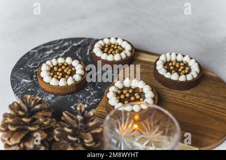 Gros plan de quatre mini-tartes de dessert au chocolat aux saupoudres dorées sur un plateau en bois et en marbre avec bougie allumée et pommes de pin décoratives. Pâtisserie festive. Tir horizontal. Photo de haute qualité Banque D'Images