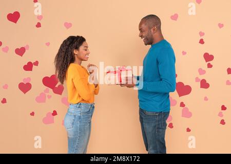 Concept d'amour. Heureux mari noir donnant cadeau à sa femme, debout sur fond pêche avec coeur volant rouge Banque D'Images