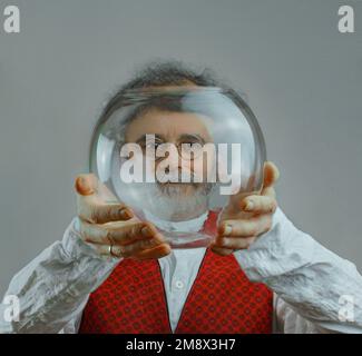 un homme coloré avec des boucles, une barbe et une moustache argentée tient une boule de verre transparente dans ses mains et regarde à travers cette balle Banque D'Images